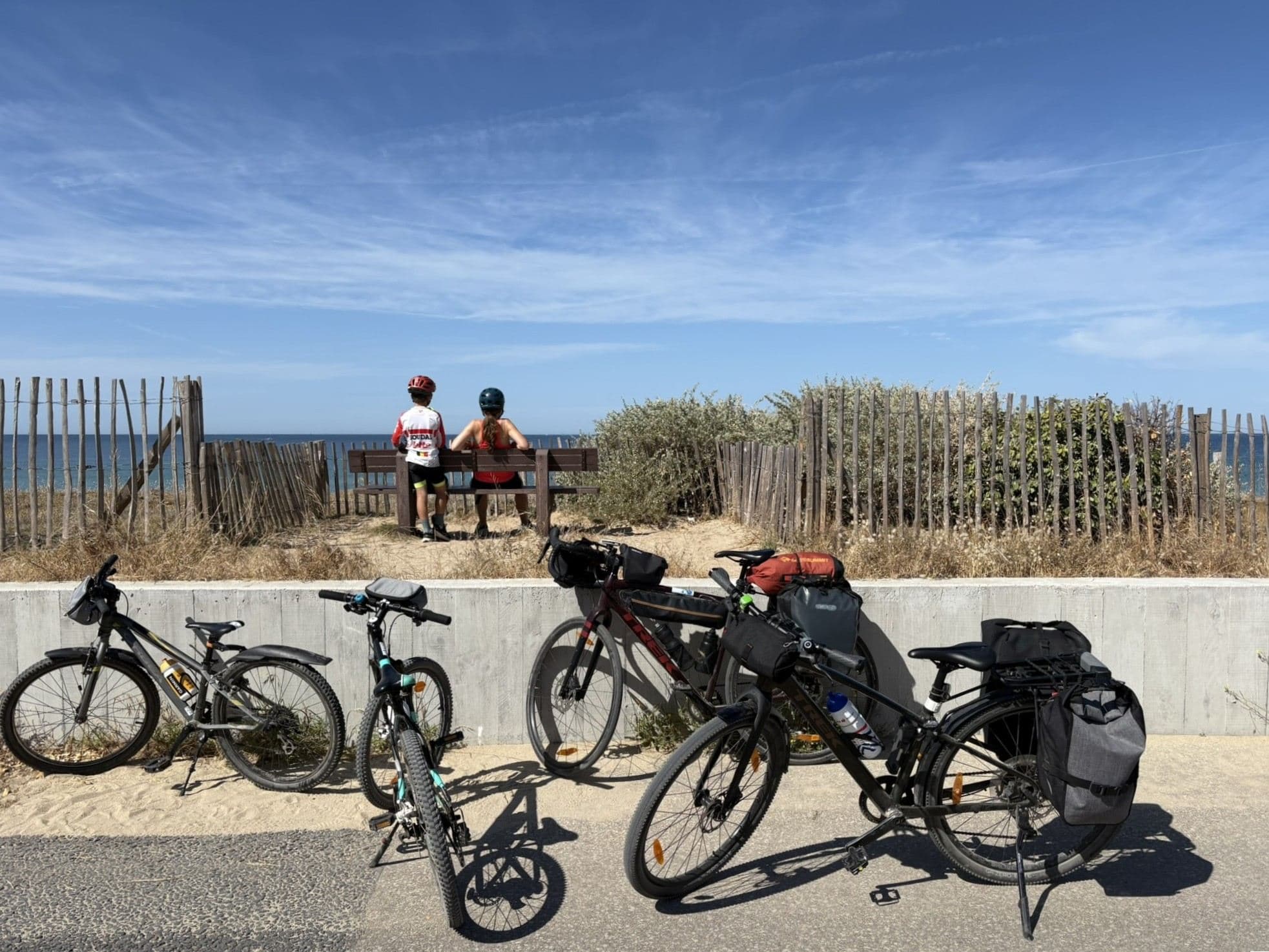 Quatre vélos chargés de sacoches contre un muret, deux cyclistes de dos face à la mer.